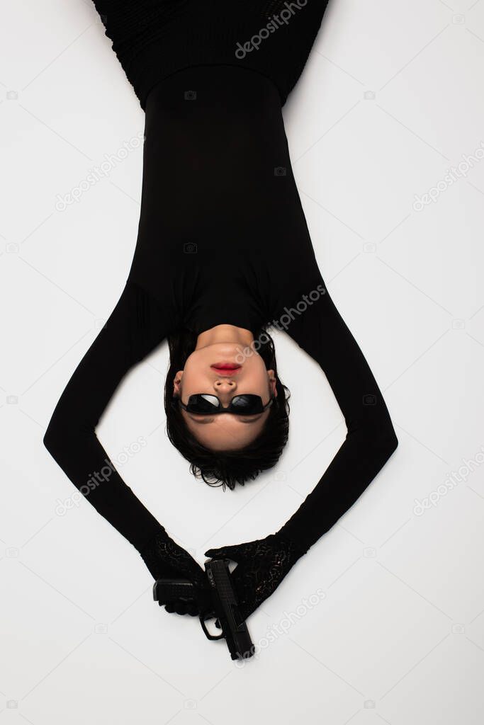 top view of brunette asian woman in black outfit and stylish sunglasses holding gun while lying on white