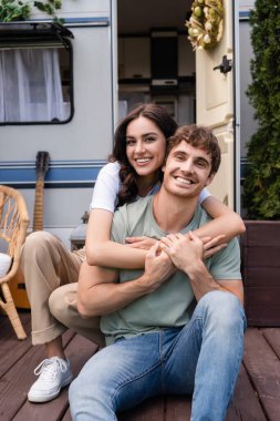 Cheerful couple hugging and looking at camera near blurred camper outdoors 