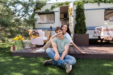 Smiling woman hugging boyfriend and looking at camera on terrace of camper van 