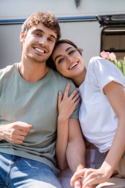 Smiling young couple looking at camera near blurred camper 