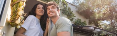 Low angle view of cheerful young couple looking away near door of camper van outdoors, banner 