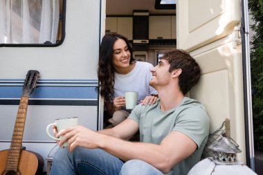 Smiling couple holding cups of coffee near acoustic guitar and camper van 