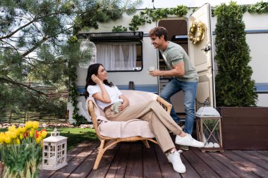 Side view of cheerful man holding cup near girlfriend on armchair and camper van 