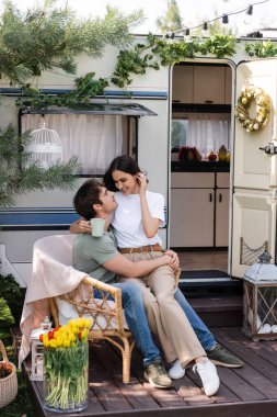 Smiling woman holding cup while sitting on boyfriend near camper van 