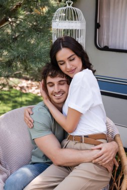 Brunette woman hugging boyfriend with closed eyes on armchair near camper van 