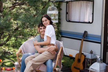 Positive woman hugging young boyfriend while sitting on armchair near camper van 