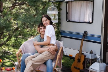 Positive woman hugging young boyfriend while sitting on armchair near camper van  clipart