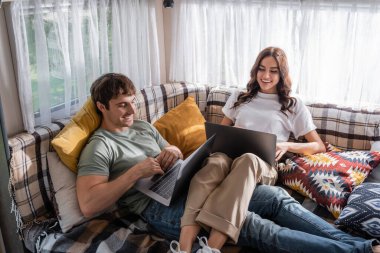 Smiling couple using laptops on bed in camper van 