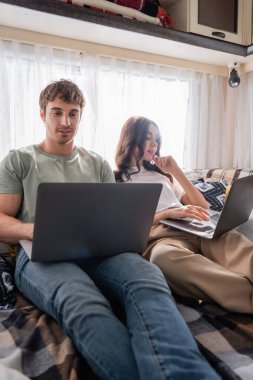 Smiling man using laptop near girlfriend on bed in camper van 