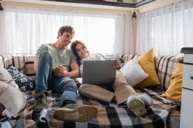 Cheerful man looking at girlfriend using laptop on bed in camper van 