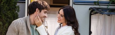Smiling woman with wine touching face of boyfriend in cardigan near camper van, banner 