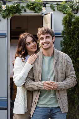 Young woman hugging boyfriend in cardigan holding wine near camper van 