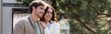 Smiling brunette woman in warm cardigans hugging near blurred camper van, banner 