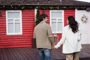 Side view of smiling couple in cardigans holding hands near blurred house 