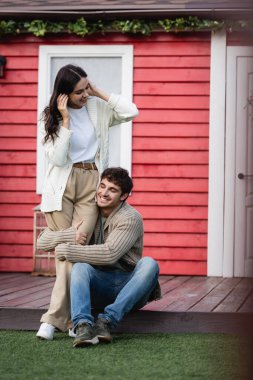 Cheerful man in cardigan hugging legs of brunette girlfriend near house 