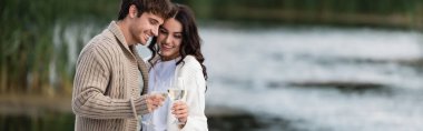 Positive couple in warm cardigans toasting wine near blurred lake, banner 