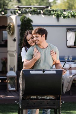 Positive woman embracing boyfriend cooking on grill near blurred camper van outdoors 