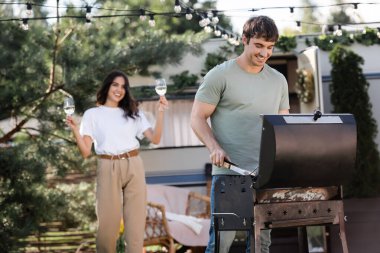 Smiling man cooking on grill near blurred girlfriend with wine and camper van 