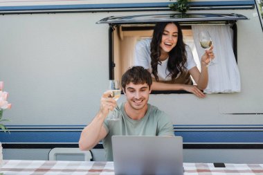 Smiling couple holding vine glasses during video call on laptop near camper van 