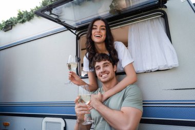 Smiling brunette woman hugging boyfriend with glass of wine near camper van 