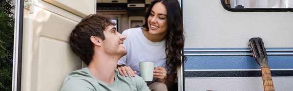 Smiling woman holding cup and hugging boyfriend near acoustic guitar and camper van, banner 