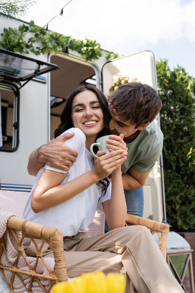 Young man kissing cheerful girlfriend with cup near camper van outdoors 