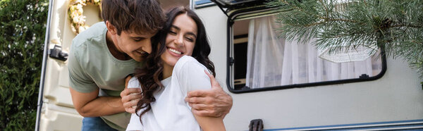 Smiling man hugging girlfriend near camper van outdoors, banner 