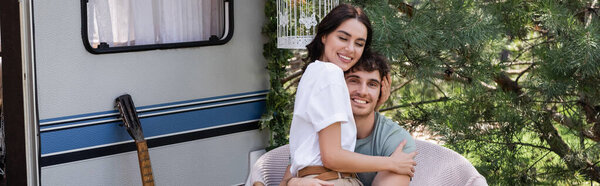 Brunette woman hugging smiling boyfriend near acoustic guitar and camper van, banner 