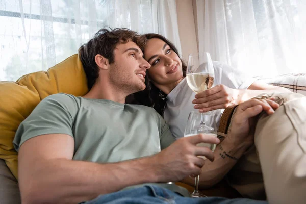 Smiling man holding glass of wine near girlfriend on bed in camper van