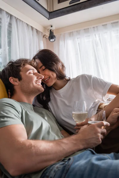 Smiling woman sitting near boyfriend with glass of wine on bed in camper van