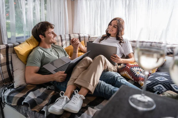 Smiling couple holding hands near laptops and glasses of wine in camper van