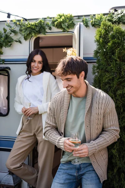 Smiling man in knitted cardigan holding wine near blurred girlfriend and camper