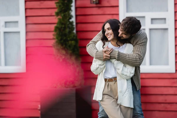 Man in warm cardigan hugging smiling girlfriend near house