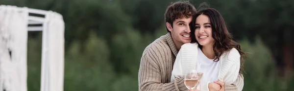 Cheerful man hugging girlfriend in cardigan holding wine outdoors, banner