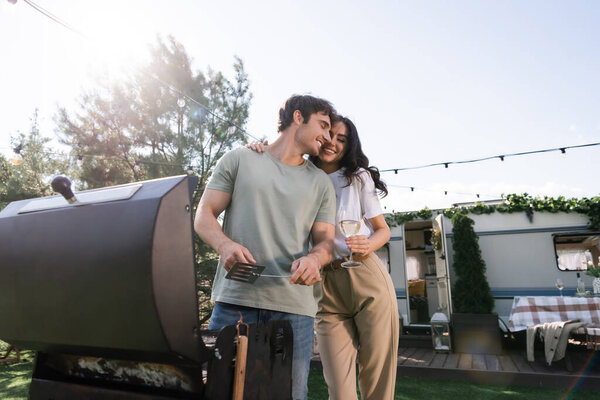 Low angle view of smiling couple holding wine and cooking near camper van 