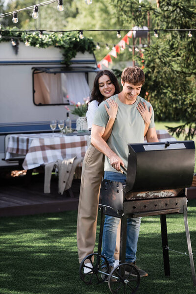 Young woman hugging boyfriend cooking on grill near blurred camper van 