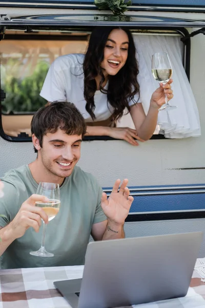 Positive couple with wine having video call on laptop near camper van