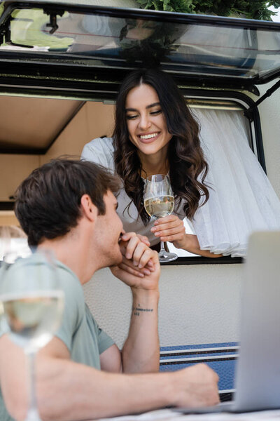 Smiling man kissing hand of girlfriend in camper van near vine and laptop outdoors 