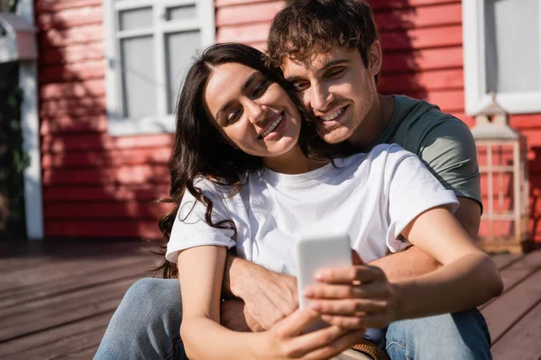 Positive man hugging girlfriend with blurred smartphone near building outdoors 