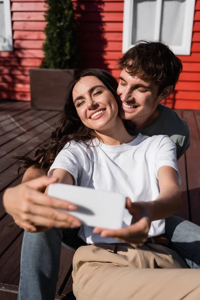 Smiling Brunette Couple Taking Selfie Smartphone Blurred House — стоковое фото