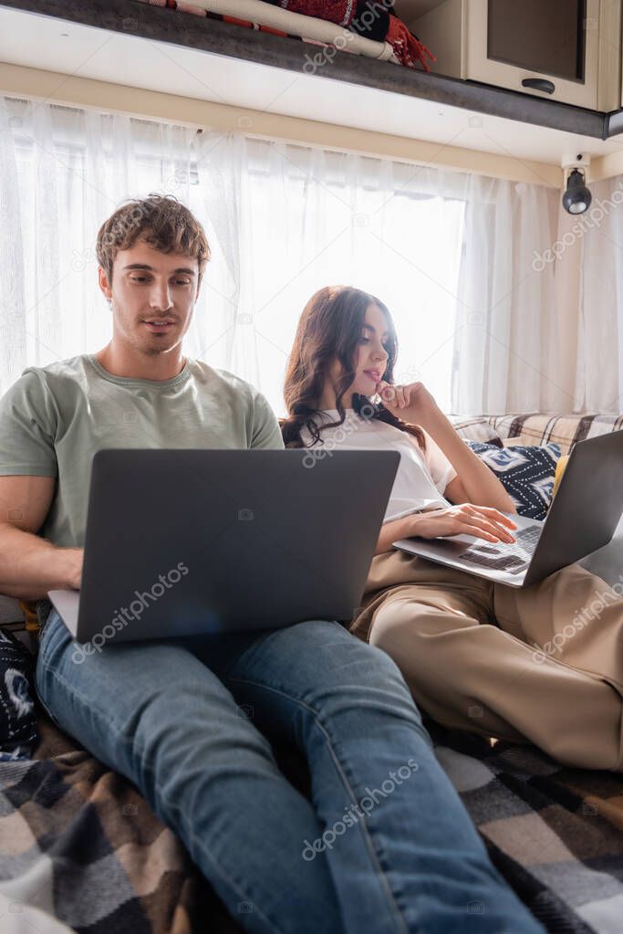 Smiling man using laptop near girlfriend on bed in camper van 