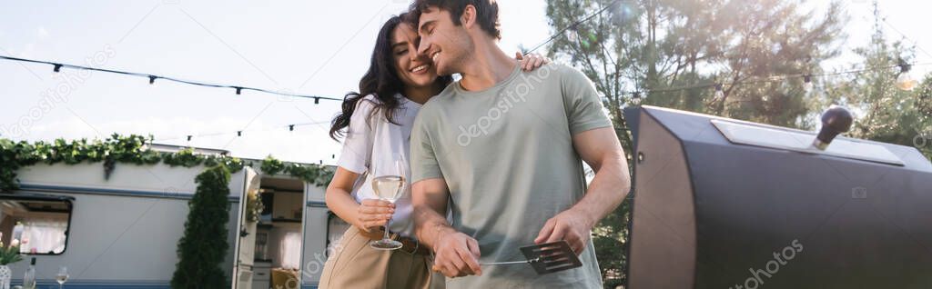 Low angle view of woman holding vine and hugging boyfriend near grill and camper van, banner 