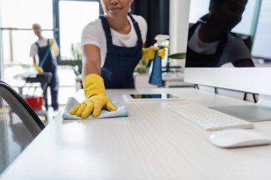 Plastik eldivenli, çift ırklı bir kadının ofis masasını temizlerken görüntüsü bulanık arka planda bir meslektaşın ve aletlerin yanında.