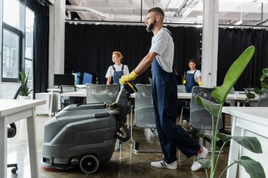 Ofiste çalışan çok ırklı temizleyicilerin yanında elektrikli süpürge makinesi olan sakallı bir adam.