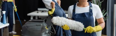 Plastik eldivenli, çift ırklı kadının arka planda, bulanık arkaplanda iş arkadaşlarının yanında toz fırçası tutarken görüntüsü.