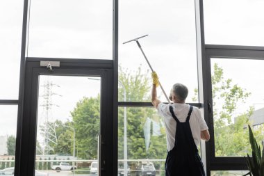 Tulumlu bir adamın pencere sileceği ile büyük ofis pencerelerini yıkamasının arka görüntüsü