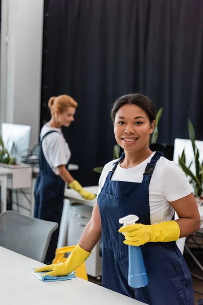 Happy Racial Woman Overalls Rubber Gloves Smiling Camera Blurred Colleague — Stock Photo, Image