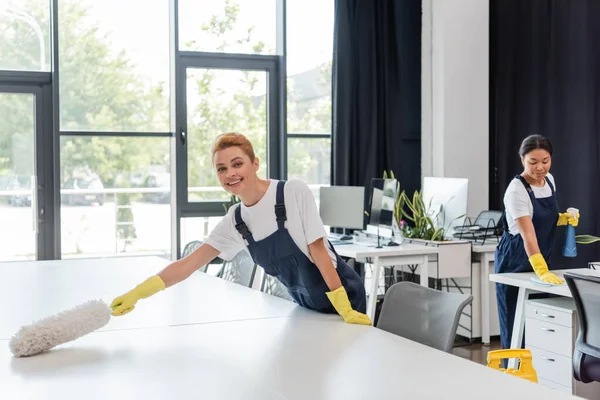 happy woman in overalls cleaning office desk with dust brush near bi-racial colleague on background