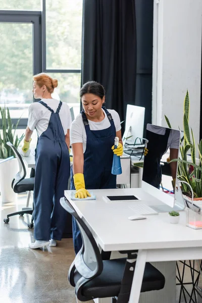multicultural women in uniform cleaning desks near computer monitors in office