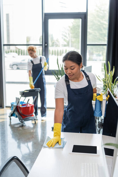 bi-racial woman washing office desk near colleague with cart of cleaning supplies on blurred background